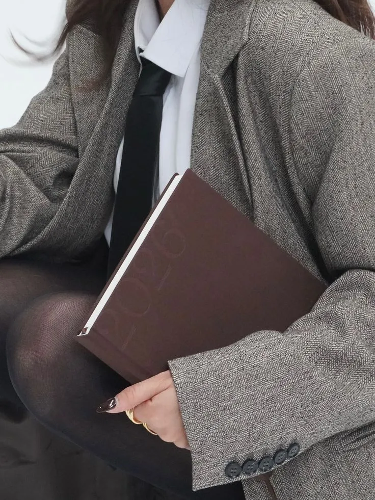 Mujer con saco gris de espiga y corbata negra sosteniendo un libro café