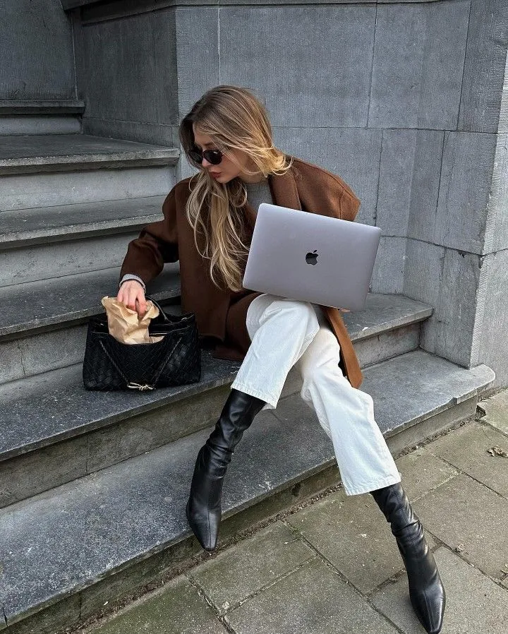 Mujer con abrigo café y laptop, sentada en unas escaleras de piedra